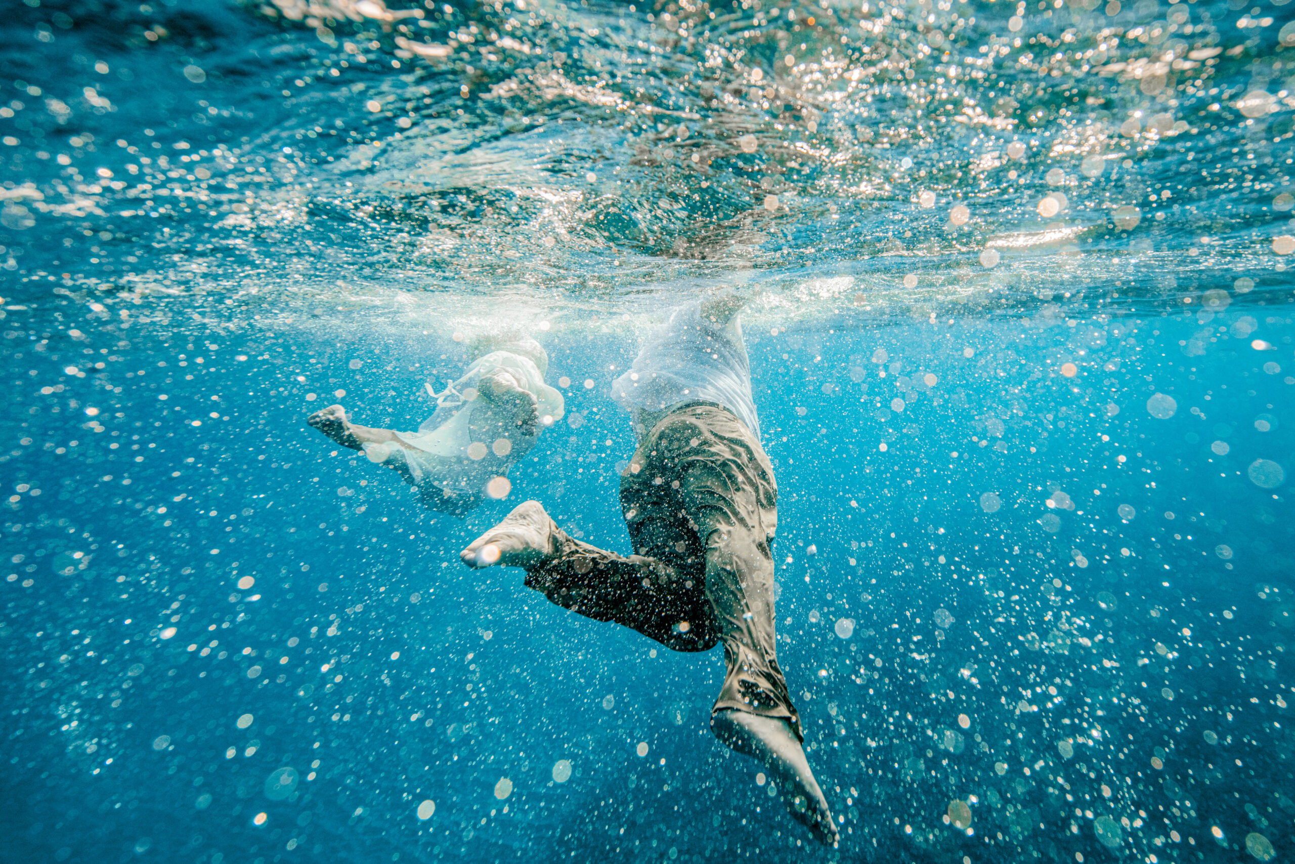 Couple is swimming underwater dramatically in their wedding clothes with bubbles all around them.